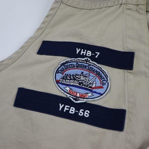 May include: Beige fabric with two navy blue patches. The top patch has the text "YHB-7" and the bottom patch has the text "YFB-56". A circular patch with a steamboat and the text "The Delta Queen Steamboat Co. Since 1890" is in the center.