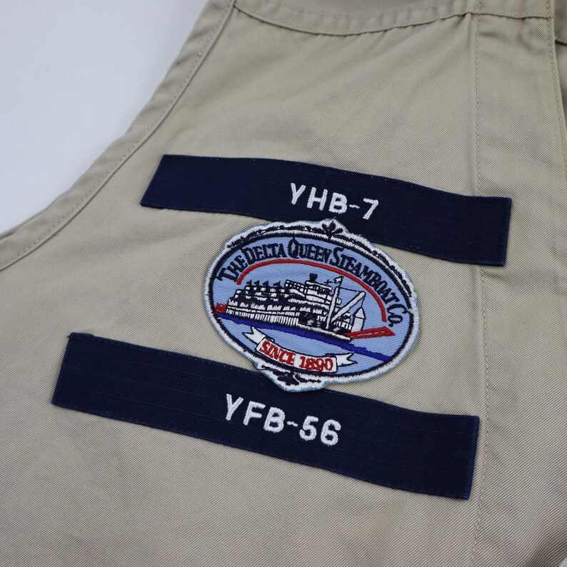 May include: Beige fabric with two navy blue patches. The top patch has the text "YHB-7" and the bottom patch has the text "YFB-56". A circular patch with a steamboat and the text "The Delta Queen Steamboat Co. Since 1890" is in the center.