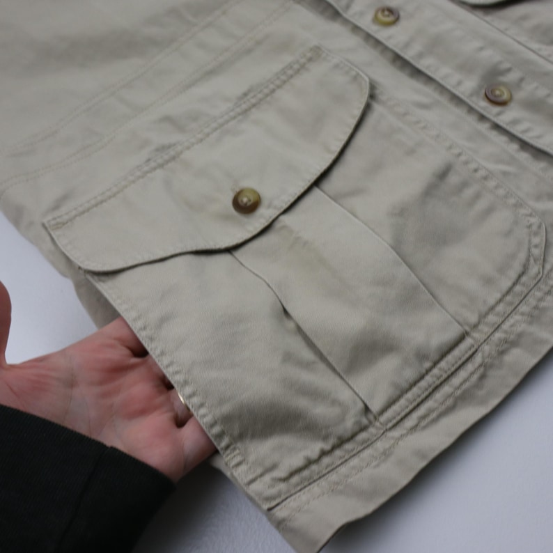 May include: A beige button-up shirt with a large, flap pocket on the front. The pocket has a button closure and a single stitch detail.
