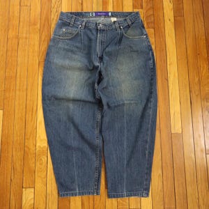May include: Blue denim jeans with a relaxed fit. The jeans have a high waist, pleats at the front, and a straight leg. The jeans have pockets and a button and zip closure. The brand name "BAGG" is visible on the waistband.
