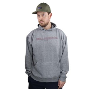Puede incluir: Una sudadera con capucha gris con un logotipo "BILLABONG" rojo y blanco impreso en la parte delantera. El modelo lleva una gorra de béisbol verde.