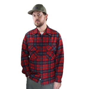 Puede incluir: Una camisa de franela a cuadros rojos y azules con dos bolsillos en el pecho. La camisa está abotonada y se usa con una gorra de béisbol verde. La gorra tiene un pequeño parche rectangular en la parte delantera.