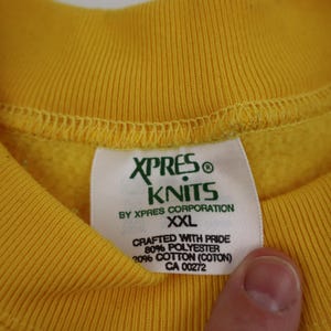 Puede incluir: Sudadera amarilla con una etiqueta verde y blanca que dice "XPRES KNITS BY XPRES CORPORATION XXL CRAFTED WITH PRIDE 80% POLYESTER 20% COTTON (COTON) CA 00272"