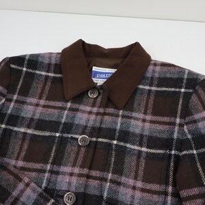 K&ouml;nnte beinhalten: Nahaufnahme einer karierten Jacke mit braunem Kragen und Kn&ouml;pfen. Die Jacke hat ein Muster aus schwarzen, braunen und lavendelfarbenen Quadraten. Der Kragen ist einfarbig braun. Die Jacke hat drei sichtbare Kn&ouml;pfe.