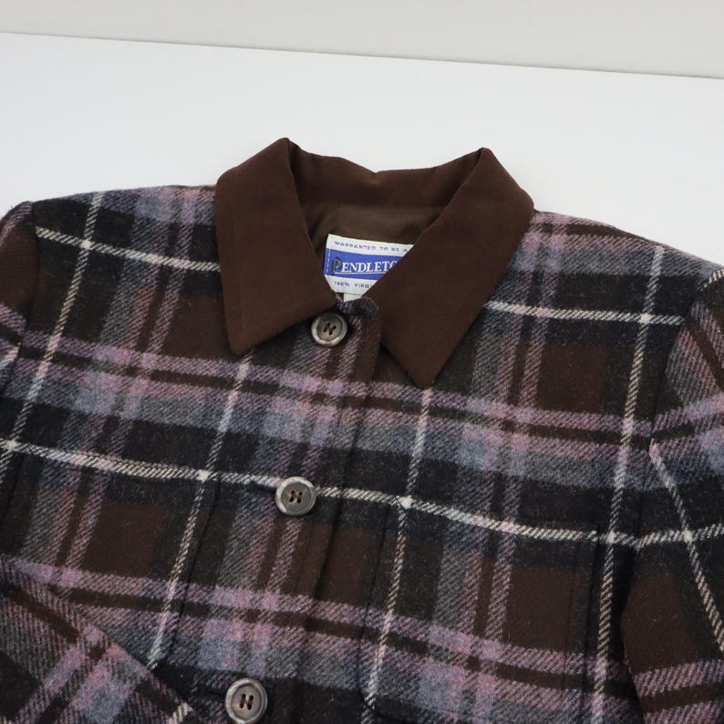 K&ouml;nnte beinhalten: Nahaufnahme einer karierten Jacke mit braunem Kragen und Kn&ouml;pfen. Die Jacke hat ein Muster aus schwarzen, braunen und lavendelfarbenen Quadraten. Der Kragen ist einfarbig braun. Die Jacke hat drei sichtbare Kn&ouml;pfe.