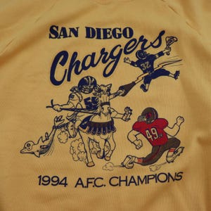 Puede incluir: Una sudadera amarilla con un logotipo vintage de los San Diego Chargers. El logotipo presenta una caricatura de un jugador de f&uacute;tbol montando a caballo, con el texto "San Diego Chargers" y "1994 A.F.C. CHAMPIONS" impreso debajo.