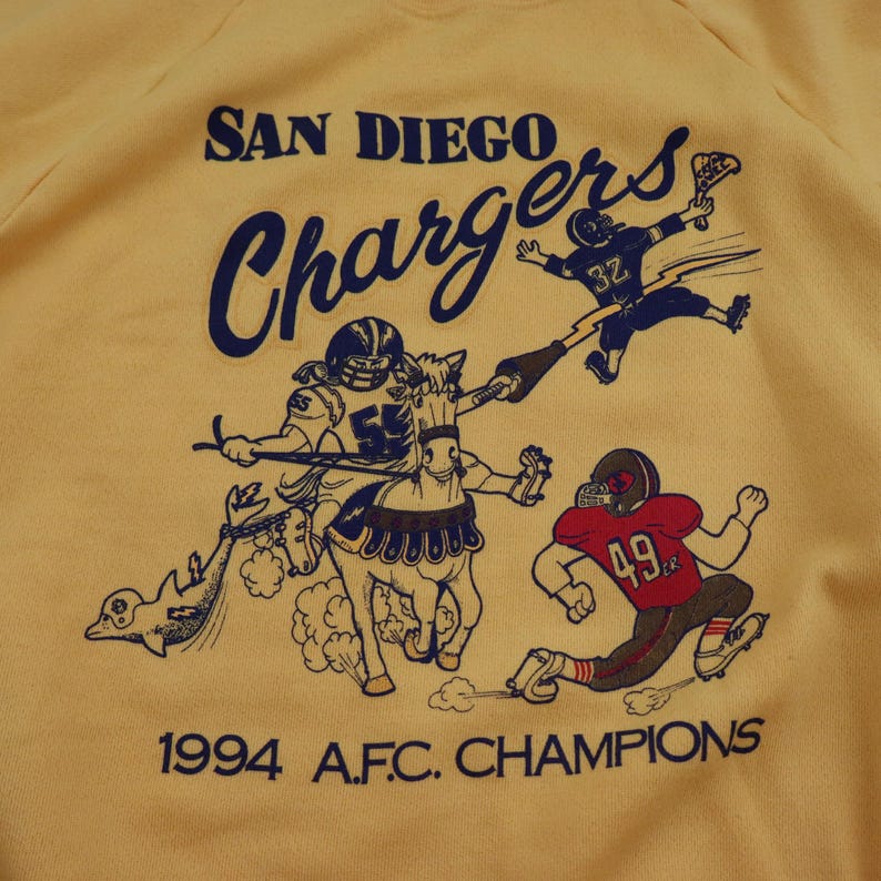 Puede incluir: Una sudadera amarilla con un logotipo vintage de los San Diego Chargers. El logotipo presenta una caricatura de un jugador de f&uacute;tbol montando a caballo, con el texto "San Diego Chargers" y "1994 A.F.C. CHAMPIONS" impreso debajo.