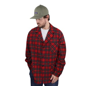 Può includere: Una camicia di flanella a quadri rossi e neri con chiusura a bottoni e una sola tasca sul petto. La camicia è indossata sopra una t-shirt bianca e abbinata a jeans blu. La persona indossa un berretto da baseball verde con un piccolo logo sul davanti.