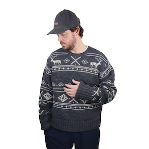 May include: A dark gray sweater with a white and gray fair isle pattern featuring reindeer and snowflakes. The sweater has a crew neck and long sleeves. The person is wearing a dark gray baseball cap.