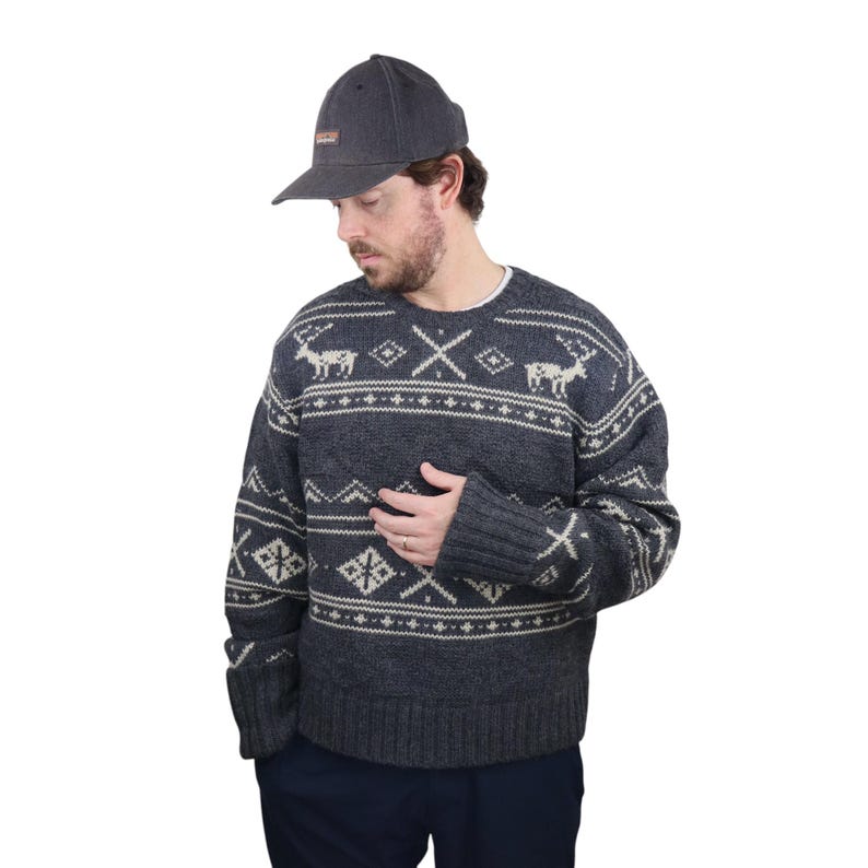 May include: A dark gray sweater with a white and gray fair isle pattern featuring reindeer and snowflakes. The sweater has a crew neck and long sleeves. The person is wearing a dark gray baseball cap.