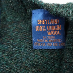Pu&ograve; includere: Primo piano di un'etichetta blu con testo dorato su un capo in lana verde. L'etichetta recita "SHETLAND 100% VIRGIN WOOL" e include testo aggiuntivo sull'origine e la cura del capo.