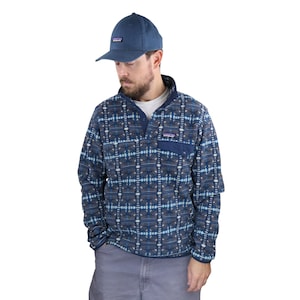 Pode incluir: Um pulôver de lã azul com um padrão geométrico em tons de azul, branco e cinza. O pulôver tem um bolso no peito e uma gola azul marinho. Um boné de beisebol azul combinando com um pequeno logotipo também é visível.