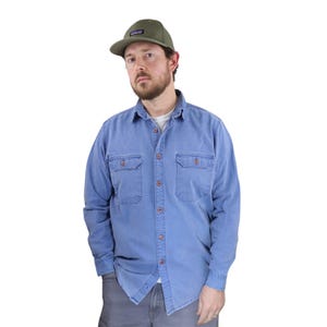 Puede incluir: Una camisa vaquera azul claro con dos bolsillos en el pecho y una botonadura. La camisa se lleva con una gorra de béisbol verde. La gorra tiene un pequeño parche rectangular en la parte delantera. La persona lleva pantalones grises.