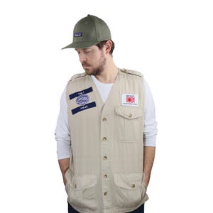 May include: A beige utility vest with multiple pockets and button closures. The vest has two patches, one with the text "YUB?" and the other with the text "DANZAI" and a Japanese flag. The person is wearing a green baseball cap with a logo.