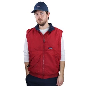 May include: A red zip-up vest with a navy blue collar and a navy blue baseball cap. The vest has a small logo patch on the chest. The cap also has a logo patch.