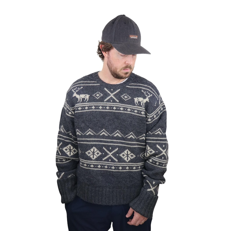 May include: A dark gray sweater with a white and gray patterned design featuring reindeer and geometric shapes. The sweater has long sleeves and a crew neck. The person is wearing a dark gray baseball cap with a small logo.