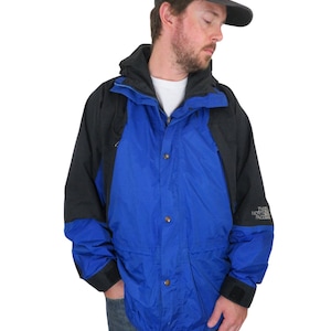 May include: A blue and black jacket with a zipper and snap closure. The jacket has a hood and a logo that says "The North Face" on the left chest.