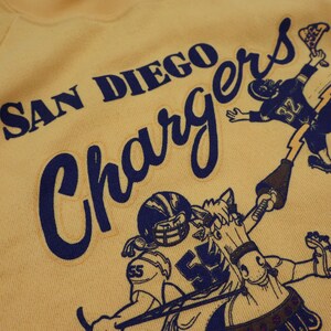 Puede incluir: Una sudadera amarilla con el texto "SAN DIEGO CHARGERS" en letras azules. La sudadera presenta una imagen de dibujos animados de un jugador de f&uacute;tbol montando a caballo.