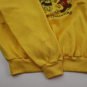 Puede incluir: Sudadera amarilla con un dibujo animado de un jugador de f&uacute;tbol y un caballero. El texto "FC CHAMPIONS" est&aacute; impreso debajo del dibujo.