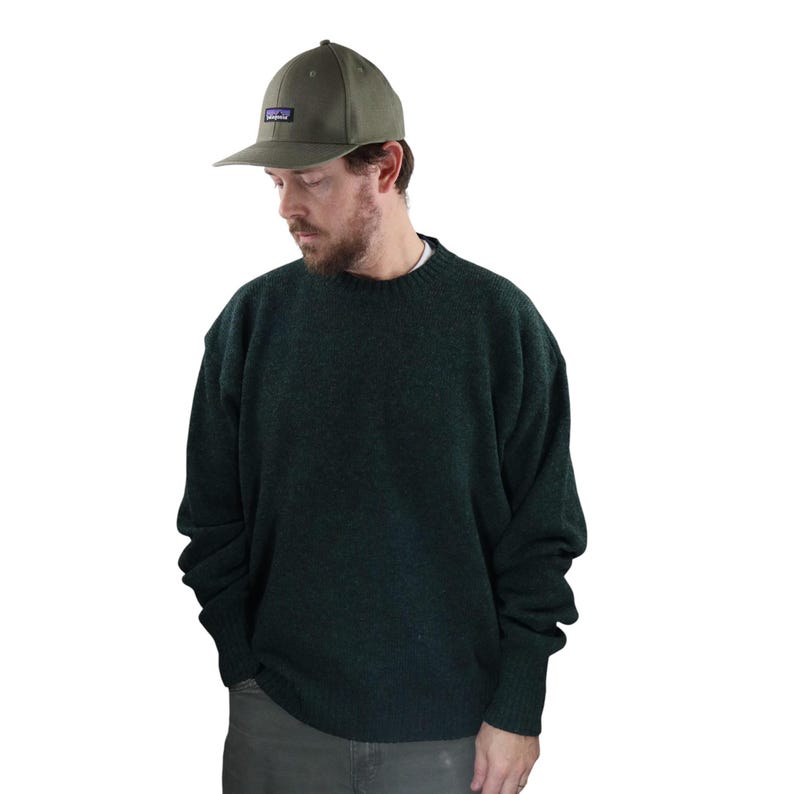 Pu&ograve; includere: Una persona indossa un maglione verde scuro e un berretto da baseball verde con una toppa rettangolare. Il berretto ha una visiera curva e una piccola toppa rettangolare con un logo. Il maglione &egrave; a tinta unita e ha uno scollo tondo.