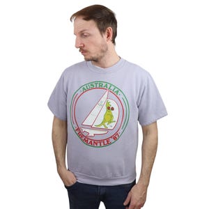Puede incluir: Una sudadera de color morado claro con un gráfico circular verde, rojo y blanco. El gráfico presenta un canguro con gafas de sol y guantes de boxeo, navegando en un barco. El texto "AUSTRALIA" y "FREMANTLE '87" está impreso alrededor del gráfico.
