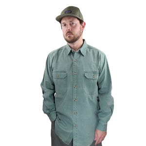 Puede incluir: Una camisa verde salvia abotonada con dos bolsillos en el pecho y una gorra de béisbol verde a juego. La gorra tiene un pequeño parche en la parte delantera. La camisa está abotonada y tiene mangas largas.