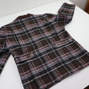 K&ouml;nnte beinhalten: Eine braune, graue und lavendelfarbene karierte Jacke mit braunem Kragen. Die Jacke hat zwei Vordertaschen mit Knopfverschl&uuml;ssen und zwei Kn&ouml;pfe vorne. Die Jacke ist aus einem gewebten Stoff gefertigt.