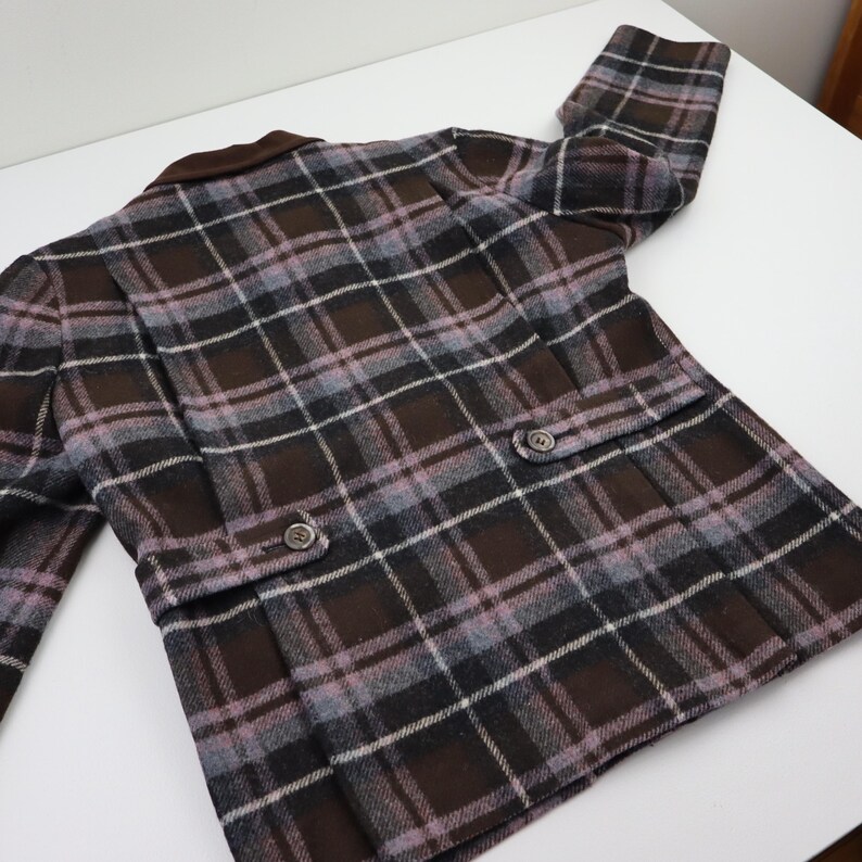 K&ouml;nnte beinhalten: Eine braune, graue und lavendelfarbene karierte Jacke mit braunem Kragen. Die Jacke hat zwei Vordertaschen mit Knopfverschl&uuml;ssen und zwei Kn&ouml;pfe vorne. Die Jacke ist aus einem gewebten Stoff gefertigt.