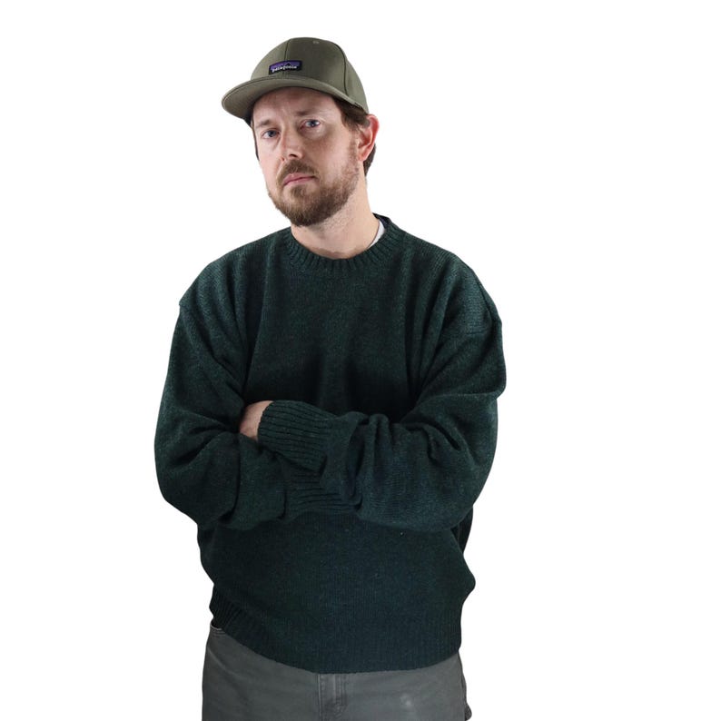 Pu&ograve; includere: Un maglione verde scuro e un berretto da baseball verde oliva. Il berretto ha una piccola toppa rettangolare sul davanti. Il maglione &egrave; a tinta unita e ha polsini a coste e girocollo. La persona indossa pantaloni grigi.