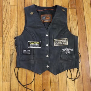 May include: Black leather vest with a V-neck, featuring patches with text and a Jack Daniel's logo. The vest has a button-up front, decorative stitching, and adjustable side laces. The patches include the phrases "CAUTION DOES NOT PLAY WELL WITH OTHERS" and "HAVING KIDS IS LIKE BEING PECKED TO DEATH BY DUCKS."