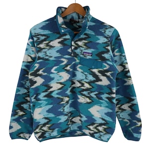 Op de afbeelding: Een blauwe, turquoise en witte fleece trui met een knoopkraag en een kleine borstzak. De fleece hangt aan een houten hanger.