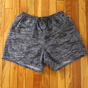 May include: Navy blue shorts with a white wave pattern and a drawstring waist. The shorts have a small purple and white logo on the lower right leg. The shorts are made of a lightweight material.