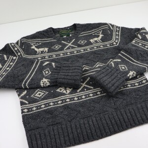 May include: A gray sweater with a white and gray patterned design. The pattern includes reindeer, trees, and geometric shapes. The sweater has a V-neck and long sleeves.