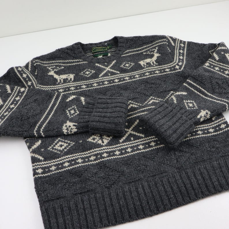 May include: A gray sweater with a white and gray patterned design. The pattern includes reindeer, trees, and geometric shapes. The sweater has a V-neck and long sleeves.
