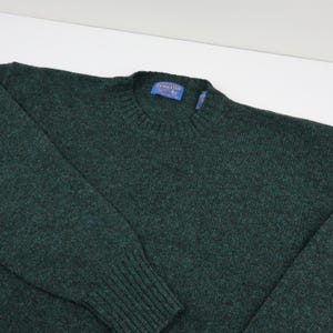 Pu&ograve; includere: Un maglione girocollo verde scuro con colletto, polsini e orlo a coste. Il maglione ha un'etichetta con la scritta "Pendleton". La maglia &egrave; realizzata in un materiale morbido e strutturato.