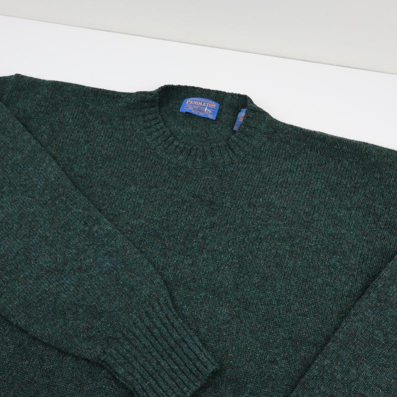 Pu&ograve; includere: Un maglione girocollo verde scuro con colletto, polsini e orlo a coste. Il maglione ha un'etichetta con la scritta "Pendleton". La maglia &egrave; realizzata in un materiale morbido e strutturato.