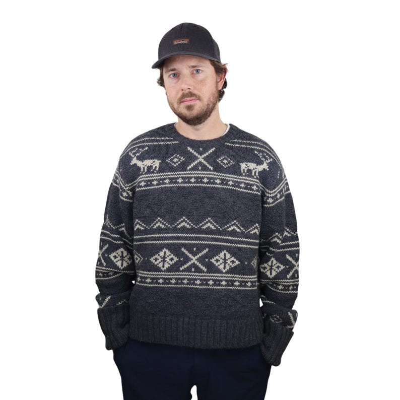 May include: A dark gray sweater with a white and gray fair isle pattern featuring reindeer and geometric designs. The sweater has a crew neck and long sleeves.