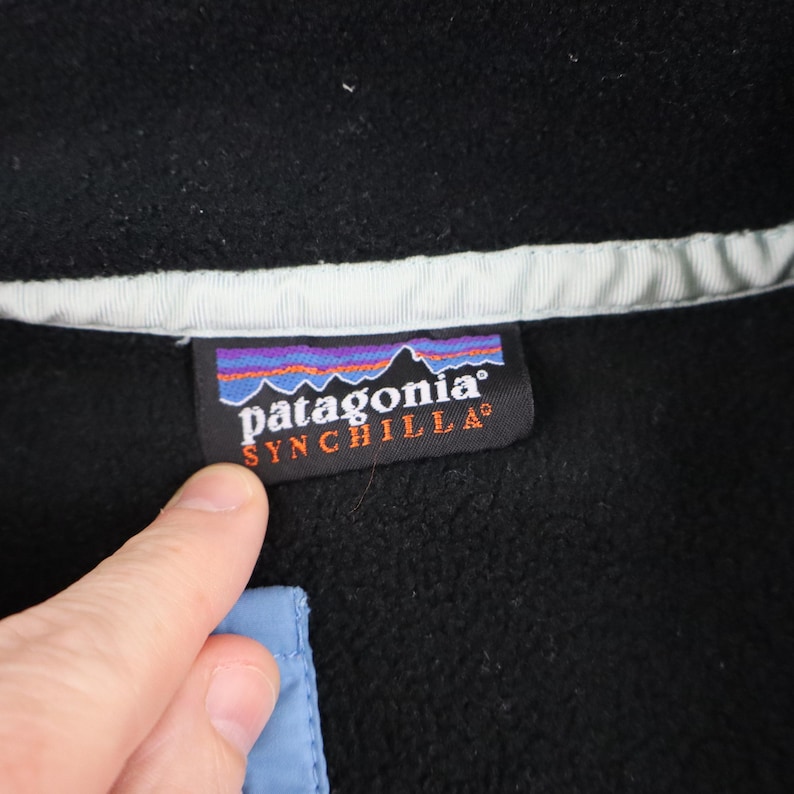 Może przedstawiać: Czarna kurtka polarowa z białym kołnierzem i jasnoniebieskim akcentem. Widoczna jest etykieta Patagonia Synchilla. Etykieta przedstawia g&oacute;rski krajobraz z nazwą marki i linią produkt&oacute;w.