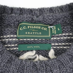 May include: A gray wool sweater with a white knit pattern. The sweater has a label that reads "C.C. Filson Co. Seattle" and "100% Wool". The label also indicates that the sweater was made in Hong Kong and is size L.