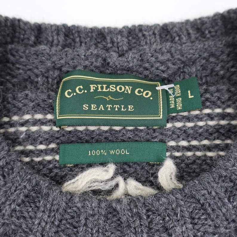 May include: A gray wool sweater with a white knit pattern. The sweater has a label that reads "C.C. Filson Co. Seattle" and "100% Wool". The label also indicates that the sweater was made in Hong Kong and is size L.