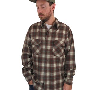 Puede incluir: Una persona con una gorra de béisbol verde con un pequeño parche rectangular y una camisa de franela a cuadros marrón, blanca y verde. La camisa tiene dos bolsillos en el pecho y está abotonada. La persona también lleva vaqueros azules.