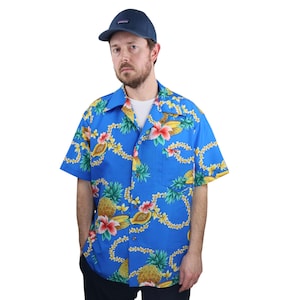 Peut inclure: Une chemise hawaïenne bleue avec un motif de fruits et de fleurs tropicales. La chemise présente des ananas, des papayes et des fleurs d'hibiscus. La personne porte une casquette de baseball bleu marine.