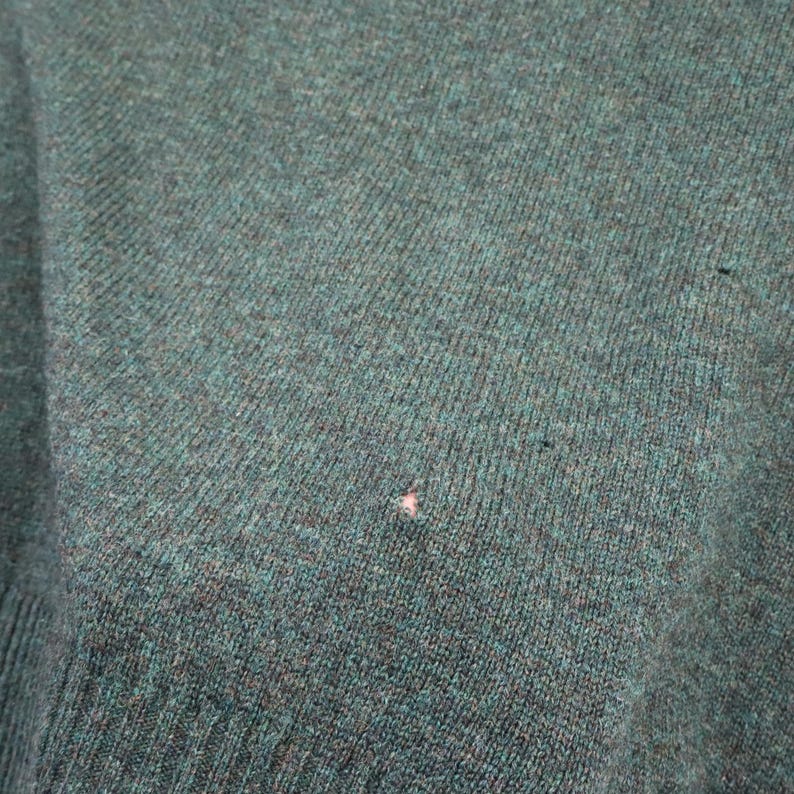 Pu&ograve; includere: Primo piano di un tessuto a maglia color verde acqua. Il tessuto ha un aspetto strutturato con linee di maglia visibili e una piccola macchia di colore chiaro. La trama generale suggerisce un maglione o un capo simile.