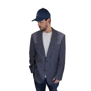 May include: A blue blazer with suede elbow patches and a two-button closure. The blazer is made of a textured fabric and has a herringbone pattern. The person is wearing a white t-shirt and blue jeans. The person is also wearing a blue baseball cap with the word "Patagonia" on the front.