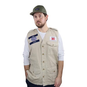 May include: A beige utility vest with multiple pockets and button closure. The vest has two patches, one with the text "YUB-7" and the other with the text "JAPANESE WILDLIFE PROTECTION", a Japanese flag, and a red sun. The person is wearing a white long-sleeve shirt and a green baseball cap.