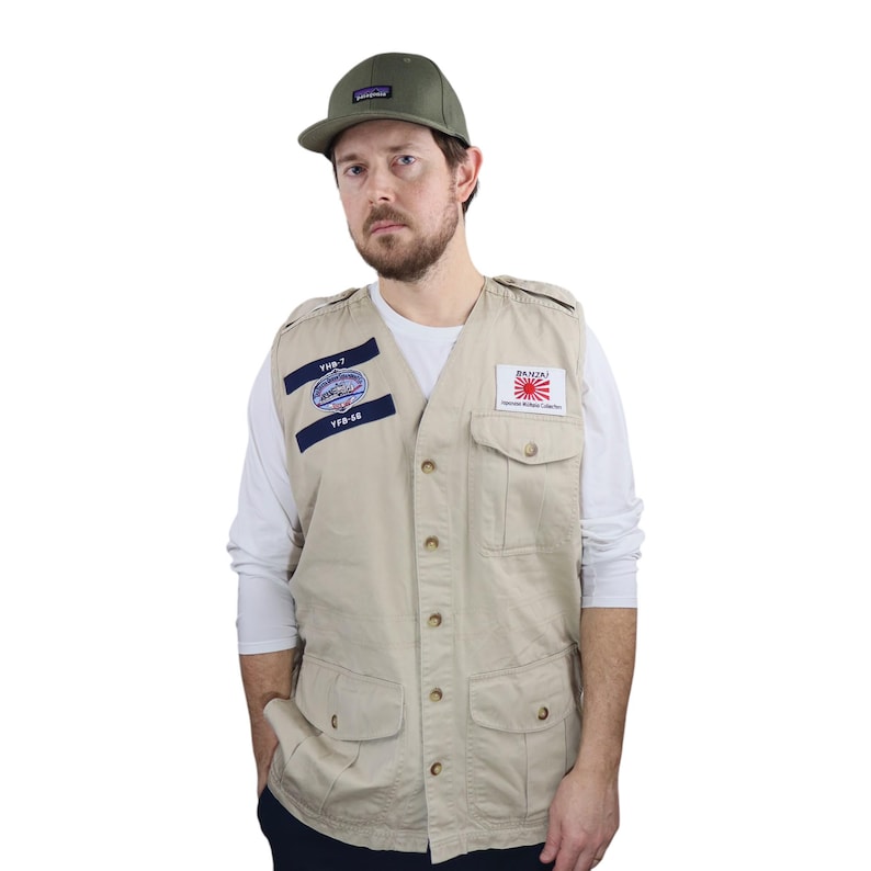 May include: A beige utility vest with multiple pockets and button closure. The vest has two patches, one with the text "YUB-7" and the other with the text "JAPANESE WILDLIFE PROTECTION", a Japanese flag, and a red sun. The person is wearing a white long-sleeve shirt and a green baseball cap.