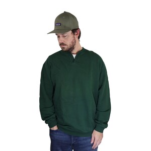 Puede incluir: Una gorra de béisbol color verde oliva con un pequeño parche rectangular con logotipo en la parte delantera, combinada con una sudadera de manga larga verde bosque oscuro con cuello con muesca y puños acanalados, sobre vaqueros azules.