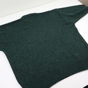 Pu&ograve; includere: Un maglione verde scuro con scollo tondo e maniche lunghe. La maglia ha orlo e polsini a coste. Il maglione &egrave; realizzato in un materiale morbido e strutturato ed &egrave; appoggiato su una superficie bianca.