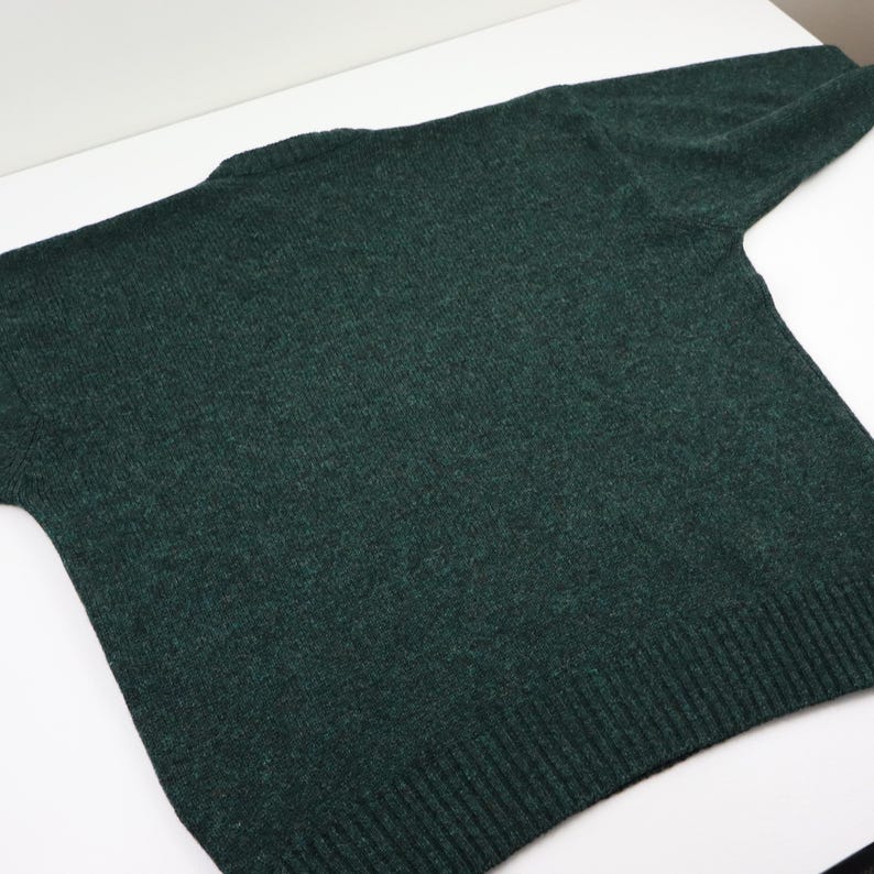 Pu&ograve; includere: Un maglione verde scuro con scollo tondo e maniche lunghe. La maglia ha orlo e polsini a coste. Il maglione &egrave; realizzato in un materiale morbido e strutturato ed &egrave; appoggiato su una superficie bianca.