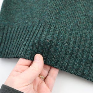 Pu&ograve; includere: Primo piano di un maglione lavorato a maglia verde scuro. Il tessuto a maglia a coste ha un aspetto strutturato. Un piccolo foro &egrave; visibile nel tessuto. Il maglione &egrave; tenuto in mano con un anello d'oro.
